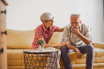 Senior couple sitting in living room. Man is sad and worried and woman is consoling him.