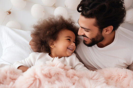 Multiracial diverse young family couple playing with cute baby child in bed holding infant kid girl. Happy multiethnic parents having fun in the morning lifting small daughter in bedroom at home