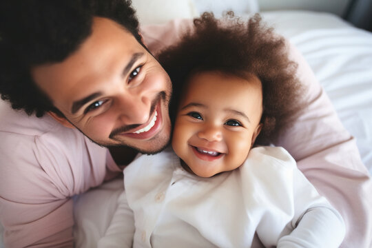 Multiracial diverse young family couple playing with cute baby child in bed holding infant kid girl. Happy multiethnic parents having fun in the morning lifting small daughter in bedroom at home