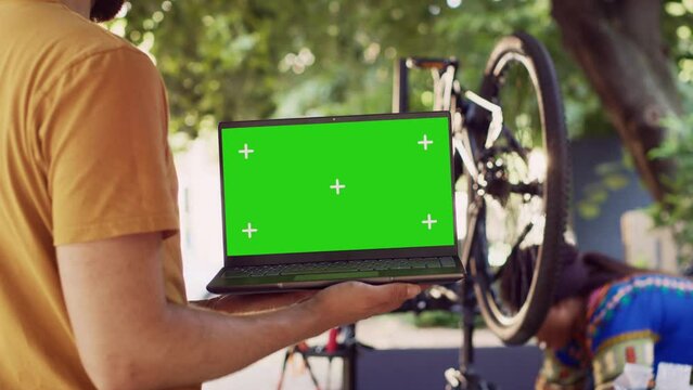 Close-up shot of laptop with isolated chromakey display carried by male cyclist while black woman dismantles damaged bicycle wheel. Couple doing bike maintenance with minicomputer having greenscreen.