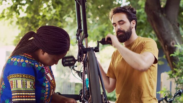 Dedicated multiracial pair cautiously fastening bike chain and pedal for summer outdoor leisure cycling. Outdoorsy young couple thoroughly examining damages on bicycle wheel for repairing.