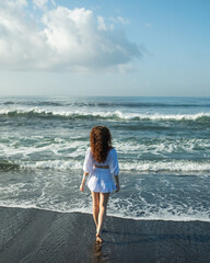 A young, beautiful girl, with curly hair, in white clothes, walks along the beach and enjoys life, rear view.