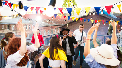 Band with guitarist, drummer and singer performing at a concert on party. Group of friends hanging out, enjoying rooftop event at sunset, dancing and listenin to music.