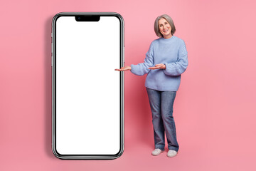 Full body size photo of promoter grandmother wearing blue knitted pullover direct hands huge phone screen new app isolated on pink color background