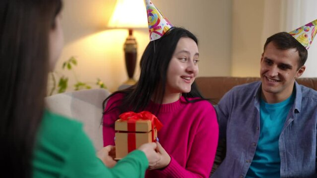 Man covering woman eyes with hands and opening as surprised lady receiving gift box from friend. Excited happy young Caucasian birthday lady enjoying gifting at home in living room