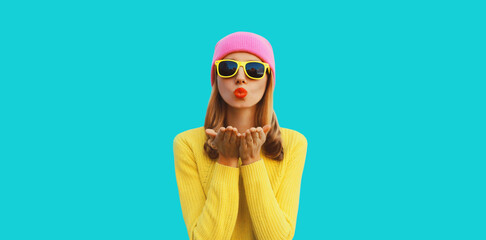 Autumn portrait of stylish beautiful young woman model blowing her lips sends sweet kiss wearing colorful hat, yellow sunglasses and sweater on blue background