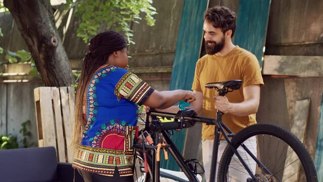 Athletic man adjusting bike on repair-stand for repairing in home yard with african american lady. Sporty caucasian boyfriend receiving help from girlfriend in realigning bicycle outside.