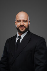 Stately bald man with a full short beard in a black suit and tie poses against a gray studio backdrop