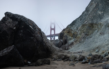 View from Marshall Beach of the Golden Gate Bridge. The bridge stands tall in the distance, partially hidden by dense fog. In the foreground, massive boulders rest on the sandy shore.
