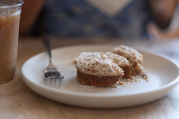 A person sits at a coffee shop, enjoying a muffin with a crumbly topping made from brown sugar and white sugar. To the side, there is a plastic cup full of creamy coffee.