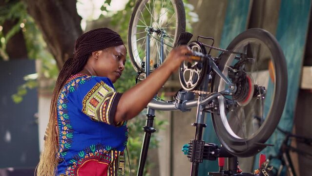 Side-view portrait of sporty healthy black woman repairing bicycle using professional tools in home yard. Female african american cyclist assessing and restoring damaged bicycle outside.