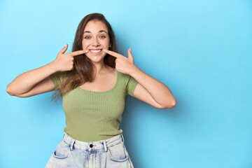 Young Caucasian woman in a green top on a blue backdrop smiles, pointing fingers at mouth.