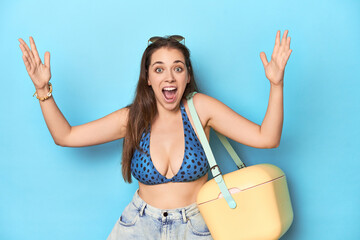 Woman in bikini with a portable beach cooler, blue studio receiving a pleasant surprise, excited and raising hands.