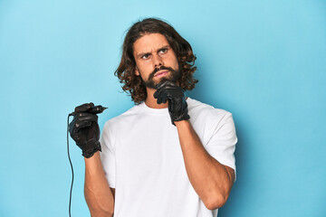 Bearded tattooist with tattoo machine at work looking sideways with doubtful and skeptical...