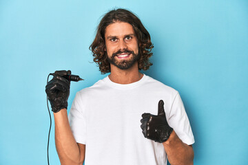 Bearded tattooist with tattoo machine at work smiling and raising thumb up
