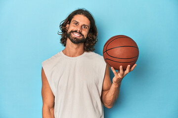 Bearded man with basketball in blue studio happy, smiling and cheerful.