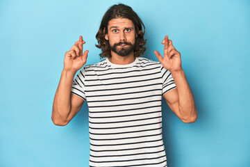 Fototapeta premium Bearded man in a striped shirt, blue backdrop crossing fingers for having luck