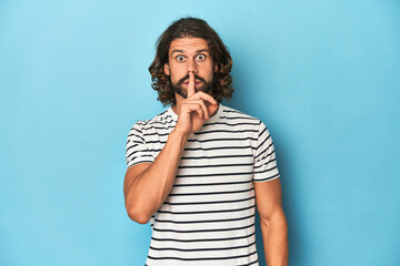Bearded man in a striped shirt, blue backdrop keeping a secret or asking for silence.