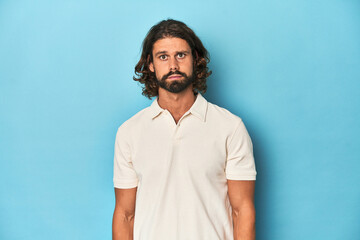 Long-haired man in a white polo, blue studio sad, serious face, feeling miserable and displeased.