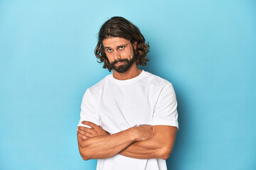 Bearded man in a white shirt, blue backdrop unhappy looking in camera with sarcastic expression.