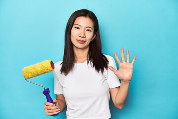 Young Asian woman with paint roller, DIY concept, smiling cheerful showing number five with fingers.