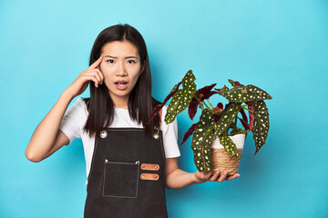 Young Asian gardener holding plant, studio backdrop, showing a disappointment gesture with...