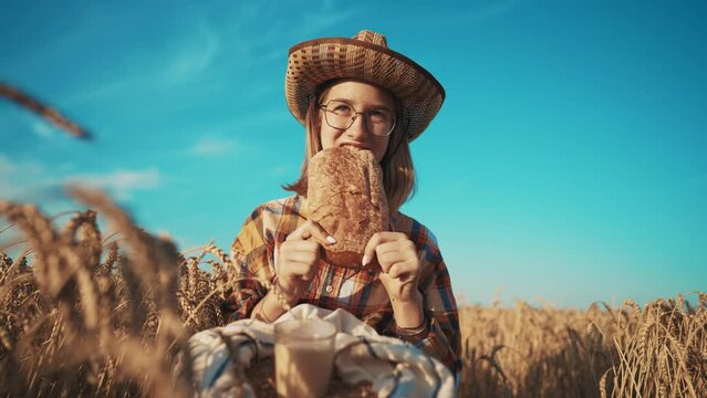 Young girl farmer in sun hat eats loaf of bread biting it drinking milk sits on wheat field with grain bag on knees. Harvesting, summer on farm, food business, agribusiness, woman worker has break.