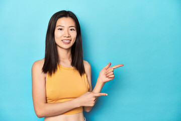 Asian woman in summer yellow top, studio setup, excited pointing with forefingers away.