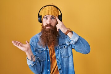 Caucasian man with long beard listening to music using headphones confused and annoyed with open palm showing copy space and pointing finger to forehead. think about it.