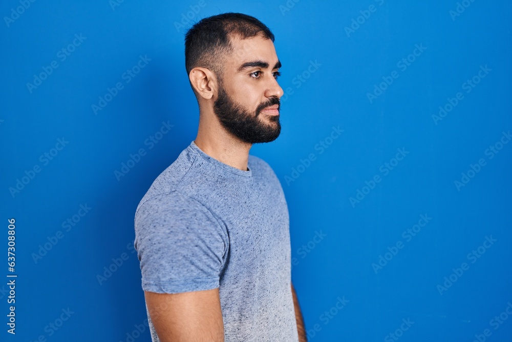 Poster Middle east man with beard standing over blue background looking to side, relax profile pose with natural face and confident smile.