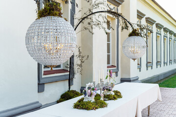 A festive table decorated with a floral arrangement with bouquets of flowers, candles and green moss in the courtyard of the estate under crystal chandeliers in the shape of a ball