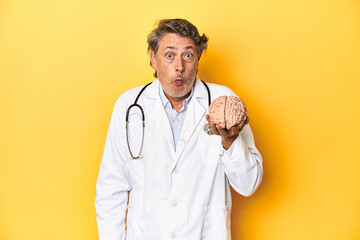 Doctor holding a brain model, yellow studio backdrop shrugs shoulders and open eyes confused.