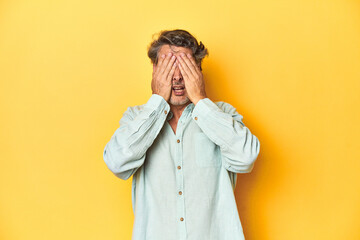 Middle-aged man posing on a yellow backdrop afraid covering eyes with hands.