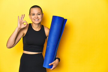 Sportswoman holding yoga mat, yellow studio background cheerful and confident showing ok gesture.