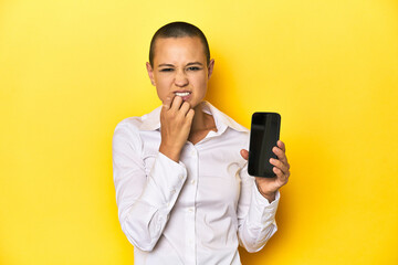 Shaved head woman holding mobile, yellow background biting fingernails, nervous and very anxious.