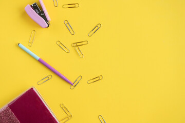 Stationery is on a yellow background. A pen and a stapler are lying next to a pink notebook