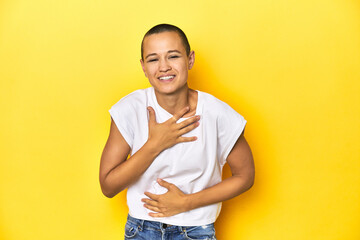 Shaved head woman in white tank top, yellow backdrop laughs happily and has fun keeping hands on...