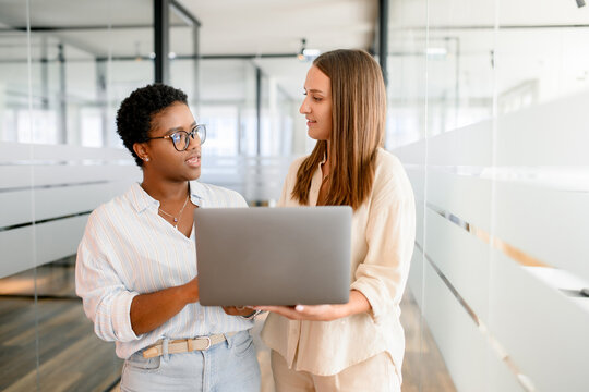 Office employees is meeting together, looking at each other in front of the laptop and discussing. Two female colleagues discussing during coffee break, coworkers have friendly conversation. Teamwork - Powered by Adobe