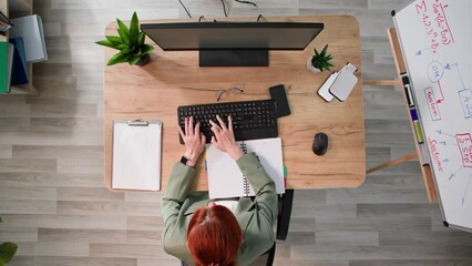 businesswoman working at computer typing on keyboard and making notes in notebook while sitting at table, top view