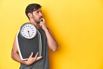 Athletic man with scale, on a yellow studio backdrop relaxed thinking about something looking at a...