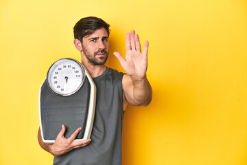 Athletic man with scale, on a yellow studio backdrop standing with outstretched hand showing stop sign, preventing you.