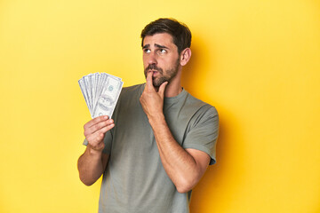 Caucasian man holding dollars, yellow studio shot relaxed thinking about something looking at a copy space.