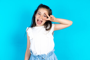 Caucasian kid girl wearing  white t-shirt over blue background Doing peace symbol with fingers over...