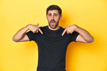 Caucasian man in black t-shirt, yellow studio backdrop surprised pointing with finger, smiling...