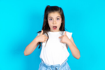 Embarrassed Caucasian kid girl wearing  white t-shirt over blue backgroindicates at herself with...