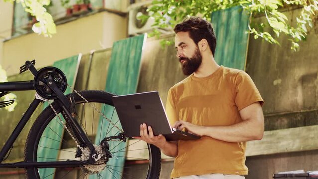 Healthy devoted caucasian man grasping his laptop to repair broken modern bicycle with online manual. Sports-loving male cyclist holding minicomputer having instructions for bike maintenance outdoor.
