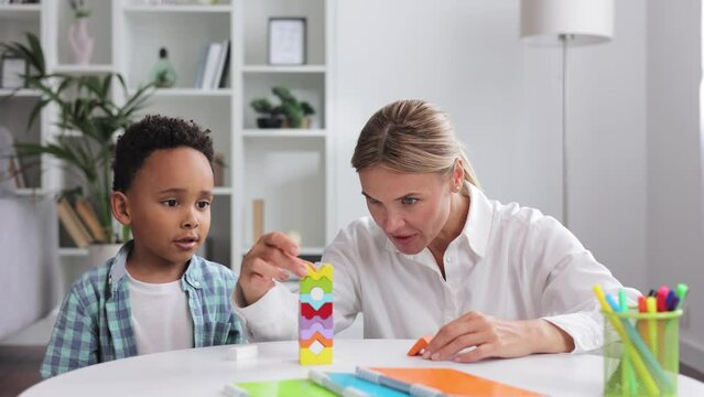 African American child curly little boy and child development specialist blonde woman playing with colorful wooden bricks sitting at table, making pyramid together. Exercises for children with autism.