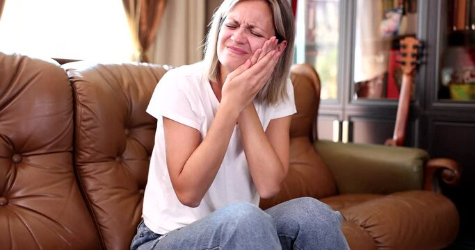 Unhealthy girl sits on the couch at home and touches cheek suffering from painful toothache. Unhappy unhealthy woman feeling unwell due to sudden severe pain in teeth