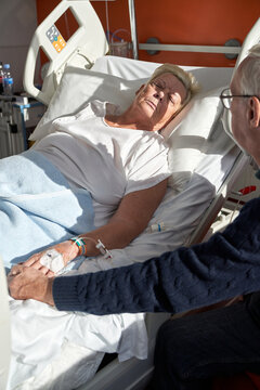 Elderly man holding hand of parient lying on bed in hospital