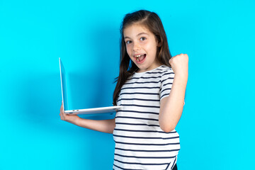 Caucasian kid girl wearing floral dress over blue background  hold computer open mouth rise fist